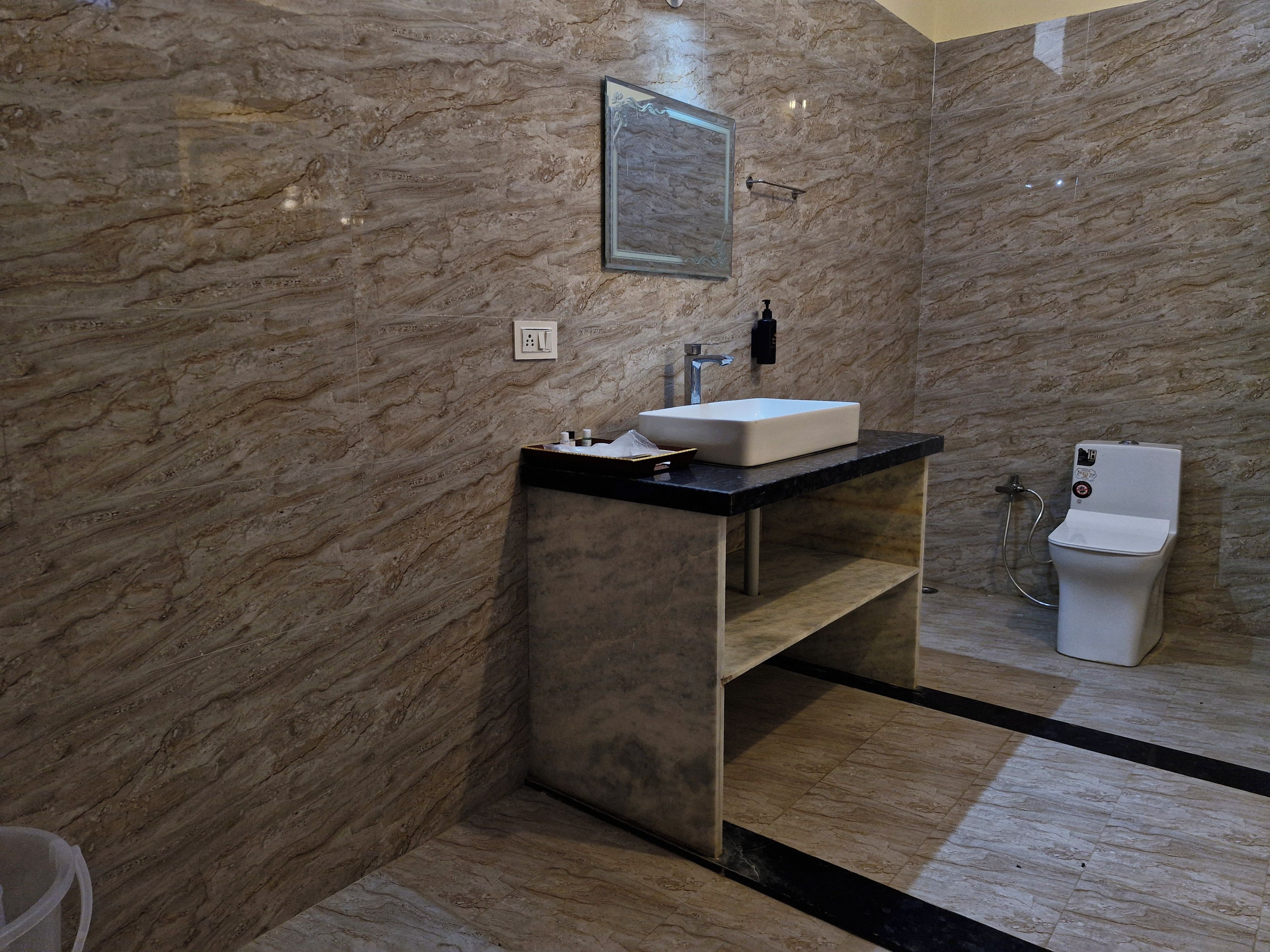 Un baño moderno con pared de mármol, un lavamanos blanco, encimera negra, inodoro y ducha.
