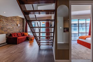 A modern interior with a brick wall, wooden flooring, a red sofa, and a staircase leading to a bedroom with a large window. - Eagle Wharf Grand Residences (London)