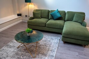 A green sofa with a blue pillow, a glass coffee table with a plant, and a patterned rug. - Eagle Wharf Grand Residences (London)