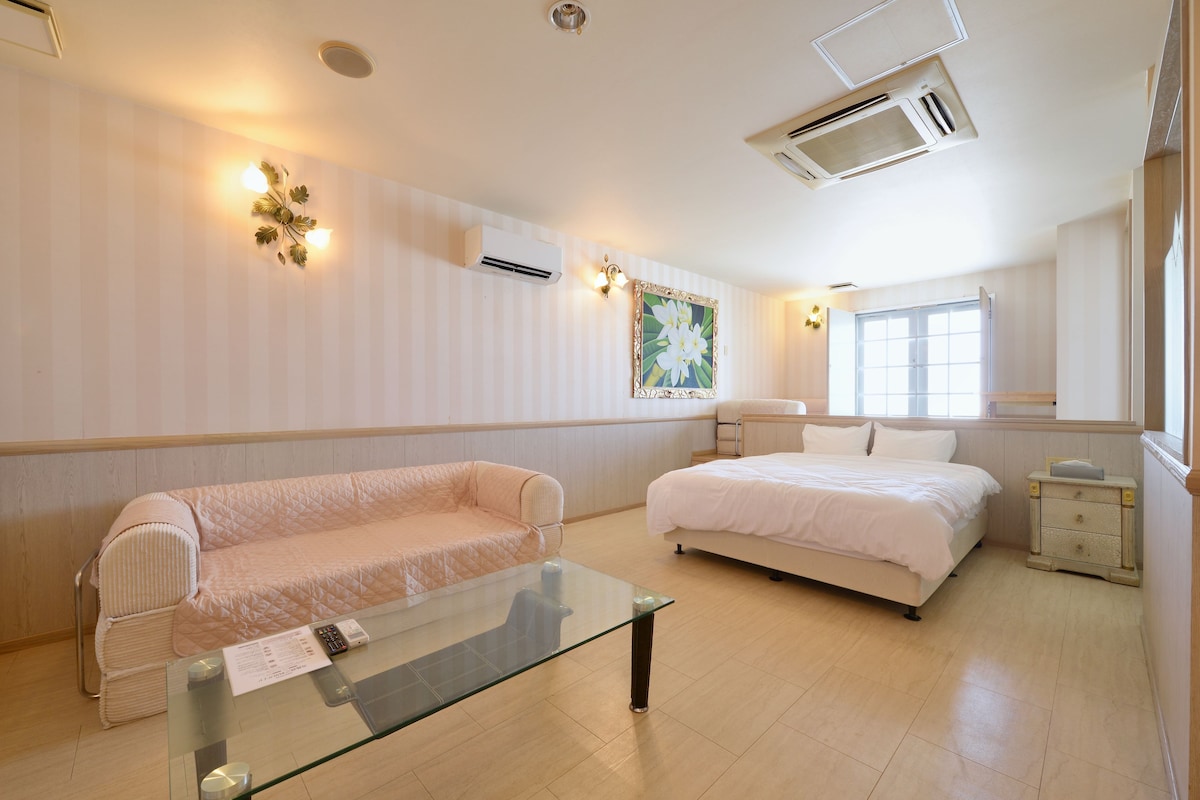 Una habitación de hotel con cama, sofá, una mesa de centro de vidrio y una mesita de noche con lámpara.