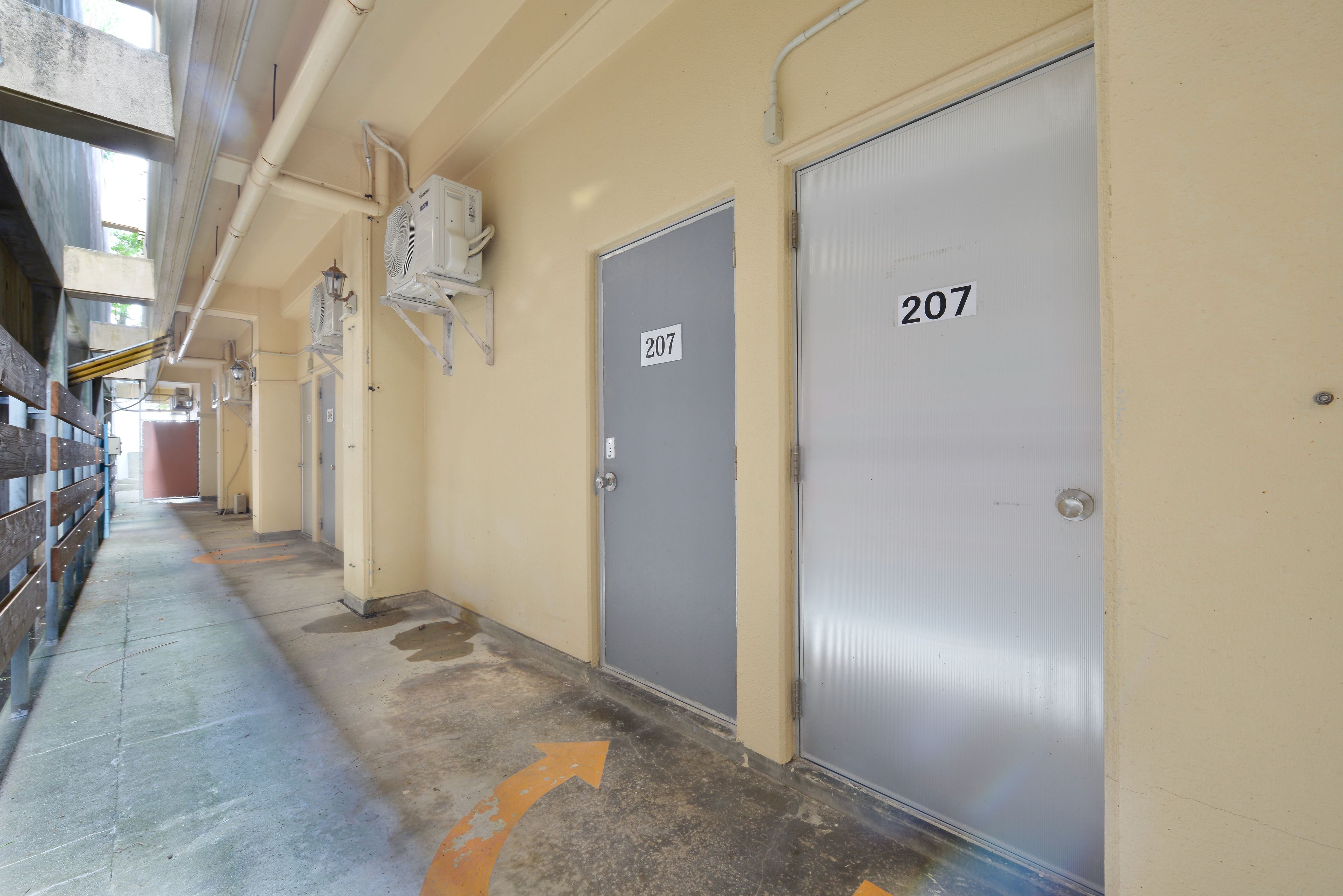A corridor with doors numbered 207 and 208, a concrete floor with a directional arrow, and overhead pipes.