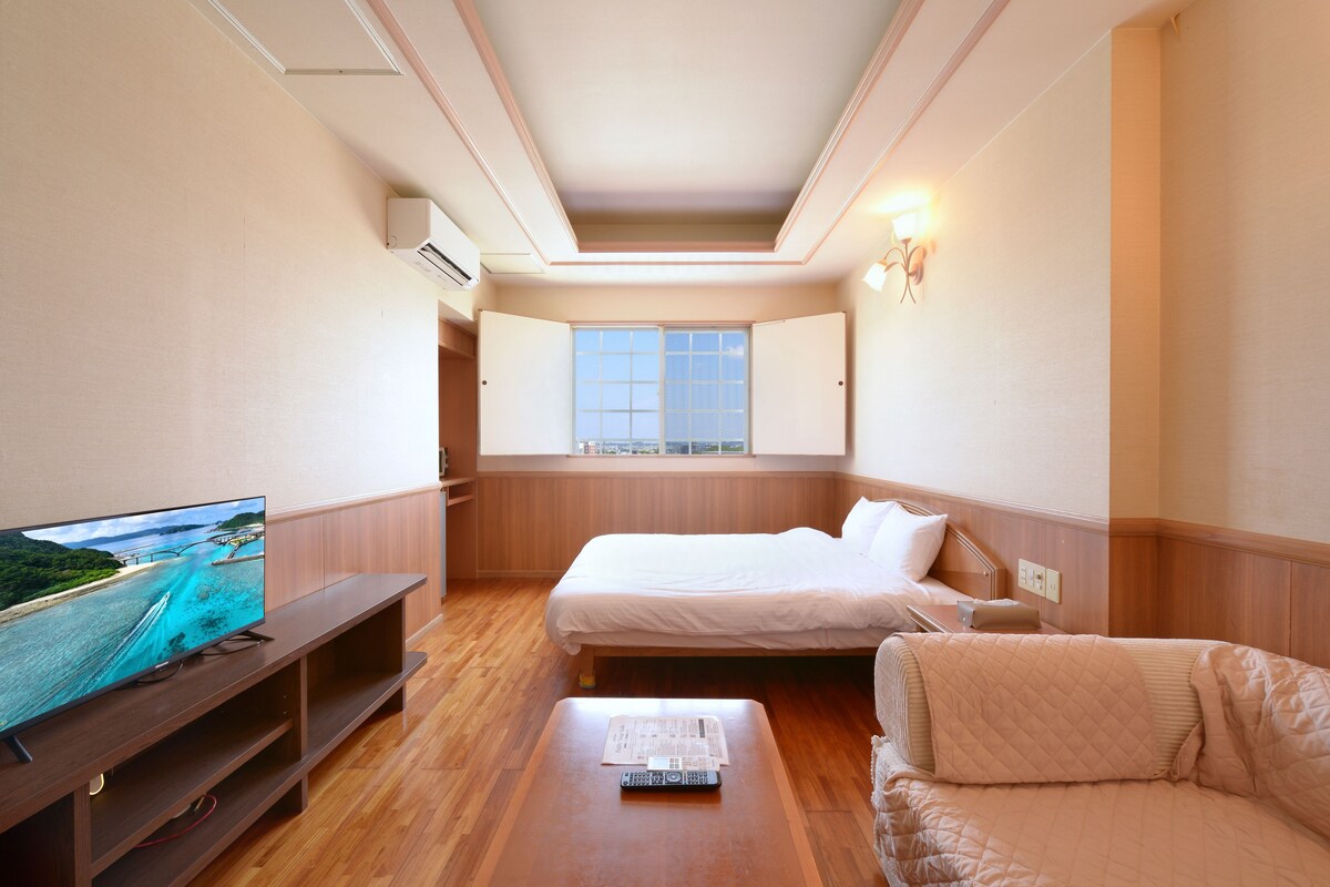 Habitación de hotel con cama, televisor sobre un soporte de madera, ventana con vista a la ciudad y mesita de noche con control remoto.