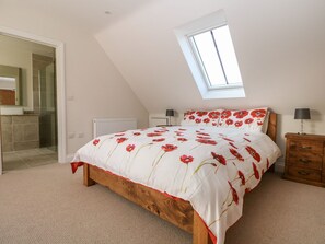 A bedroom with a sloped ceiling, a skylight, a bed with a floral bedspread, a bedside table with a lamp, and a wooden nightstand. - The Granary (Bordon)