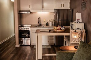 A compact kitchen with a stove, oven, microwave, and refrigerator. There is a wooden table with a placemat and a tray of snacks. A wicker chair with a light fixture is visible. - Cozy, newly renovated, one bedroom apartment in Kingston,  TN (Kingston)