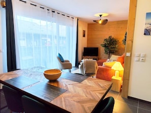 A modern living room with a television, seating area, and a dining table with a bowl. - L'Alpin (4 People) Balcony with a view of the mountains—close to the train station and the gondola. Indoor parking. (Saint-Gervais-les-Bains)