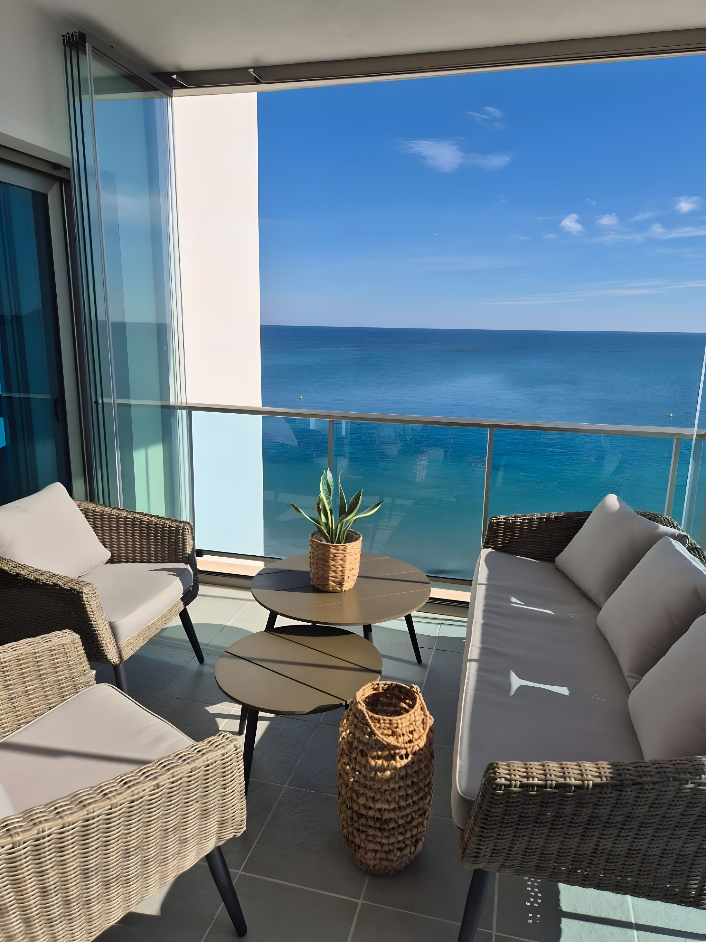 A balcony with wicker furniture, a small table, and a potted plant, overlooking a clear blue sea.