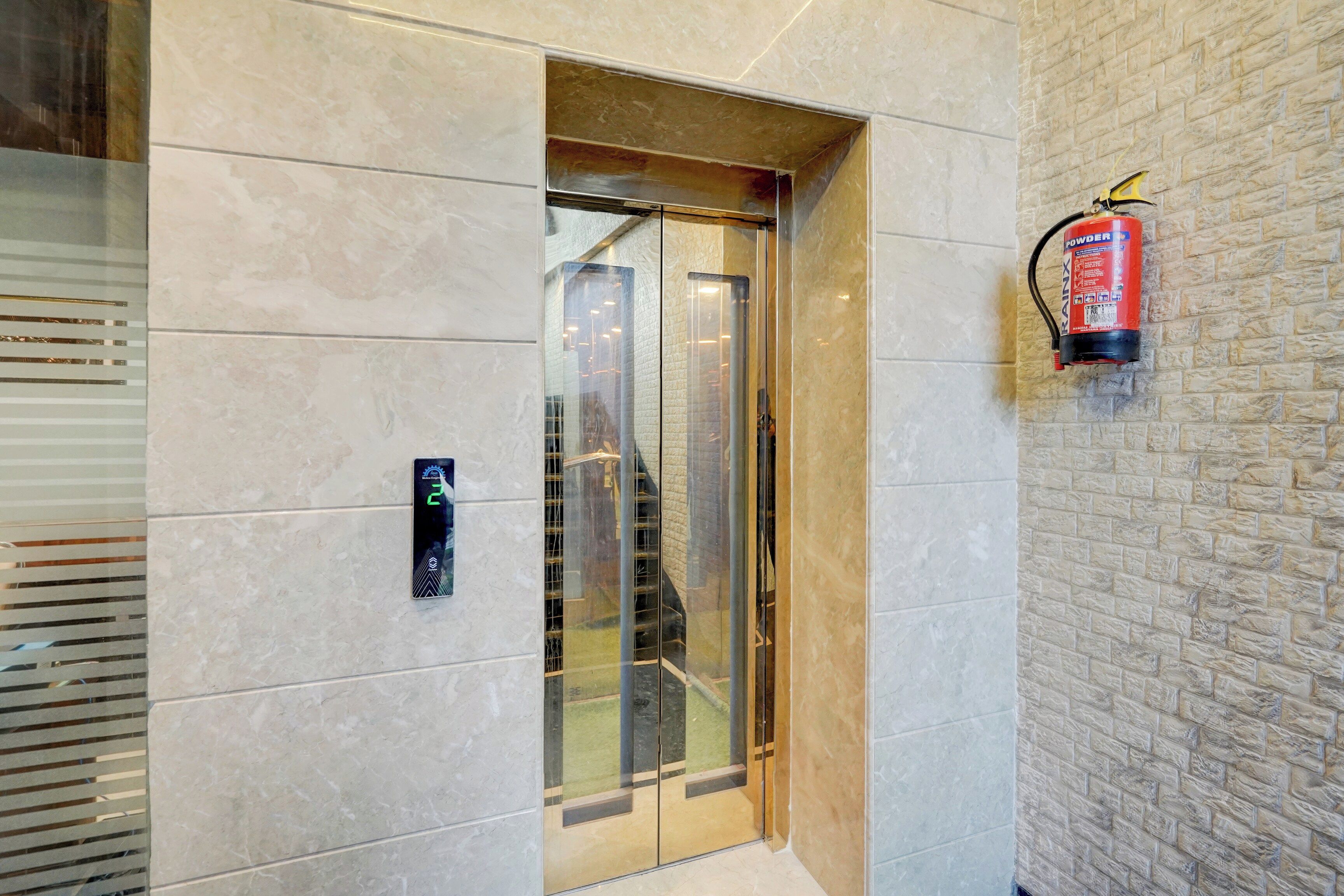 A modern elevator with a glass door, a fire extinguisher mounted on the wall, and a digital display panel on the wall.