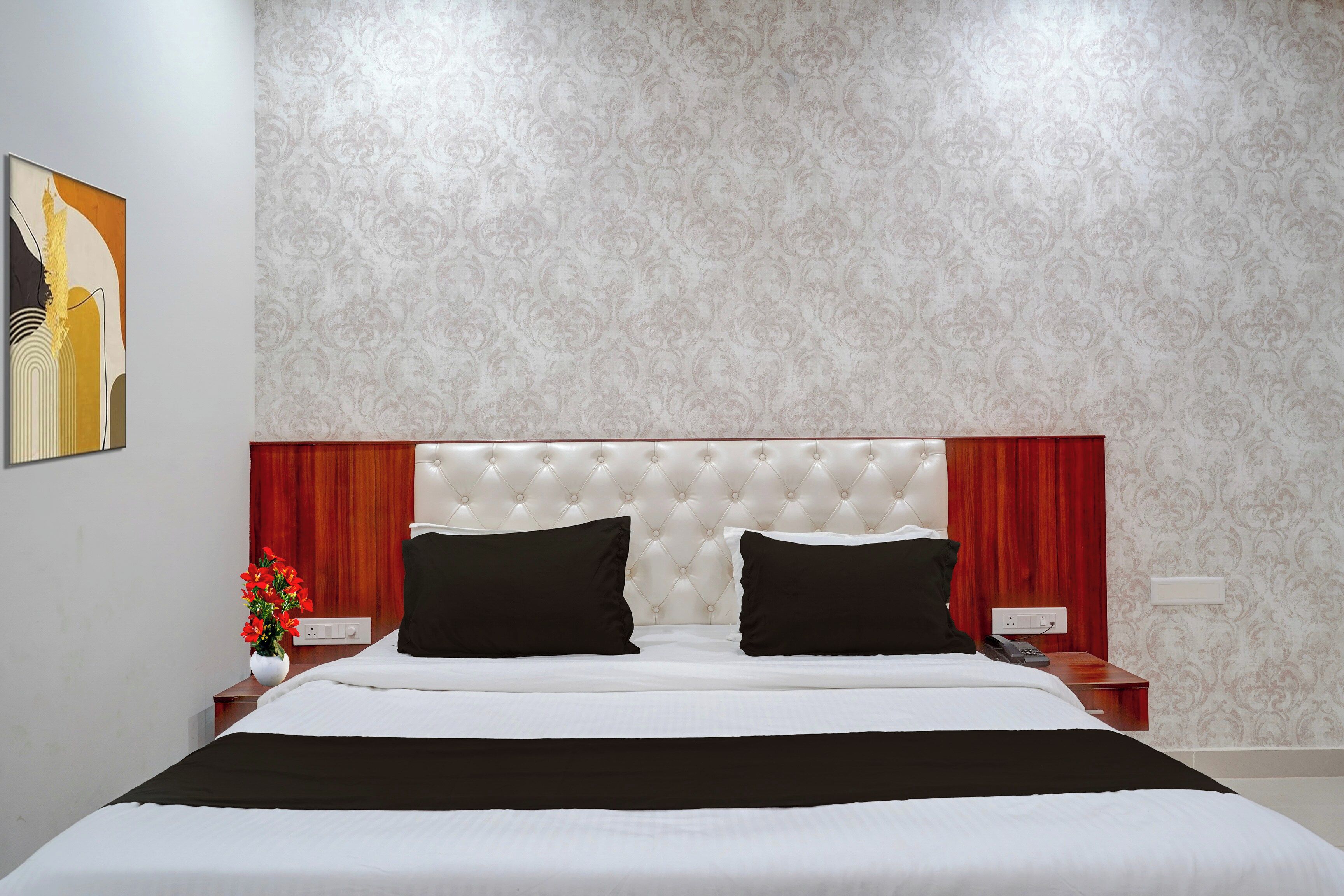 A hotel room with a tufted headboard, a bed with white linens and black accents, a nightstand with a telephone, and a wall-mounted power outlet.