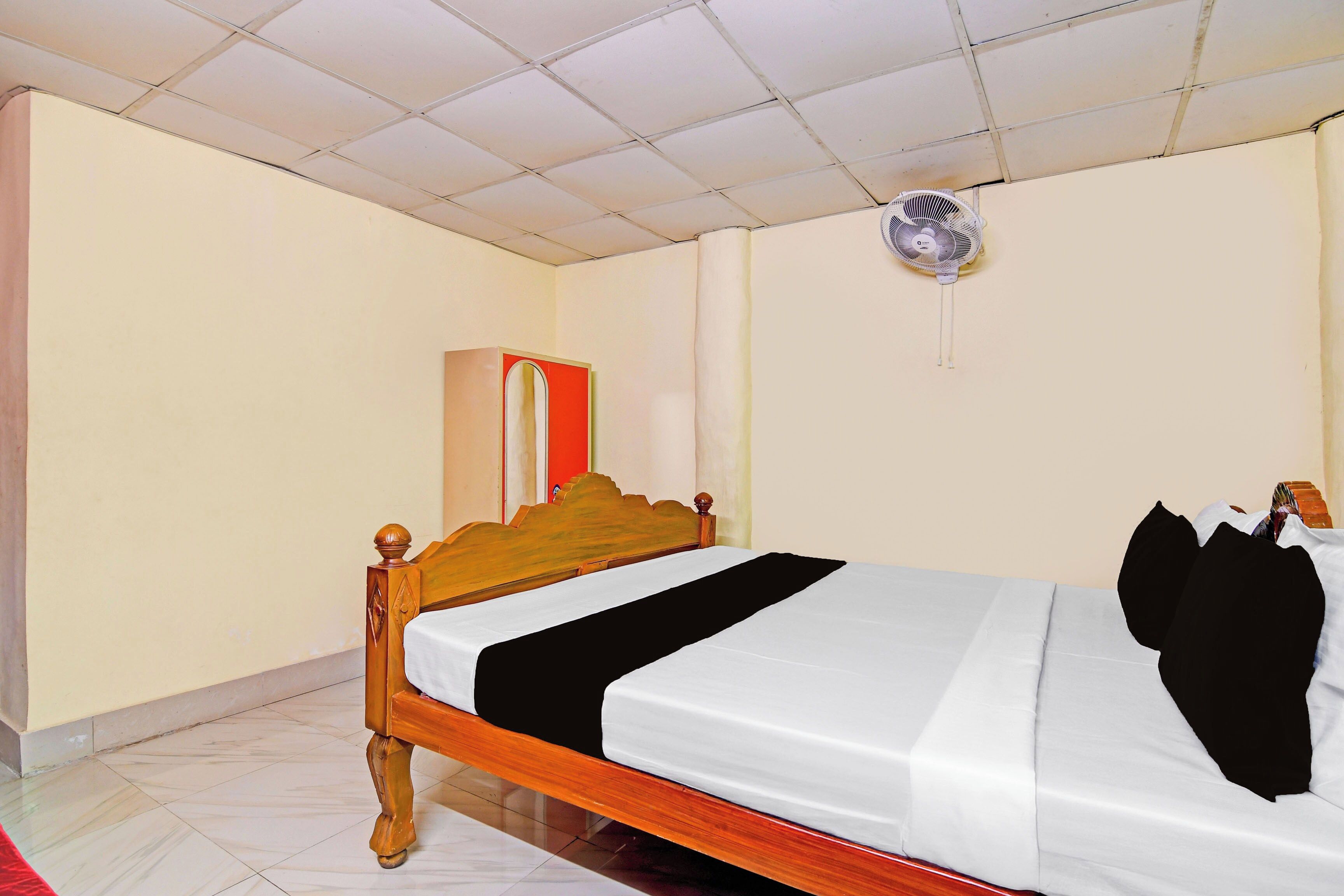 Una habitación de hotel con una cama de madera, una colcha blanca, detalles en negro, un ventilador de pie y un armario rojo.