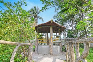 A gazebo with a wooden roof and a concrete base, surrounded by a wooden fence and lush greenery.