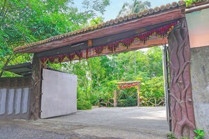 A gated entrance with a wooden archway adorned with hanging flowers, leading to a paved area with a concrete wall and a metal gate.