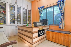 A reception desk with a sign reading 'HOTEL & Company Serviced', a window with patterned curtains, and a cabinet with glass doors containing various items.