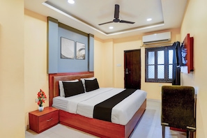 A bedroom with a bed, bedside table, wall art, ceiling fan, air conditioning unit, window with mesh screen, and a chair. - Super OYO Collection O BHU Road Varanasi (Varanasi)