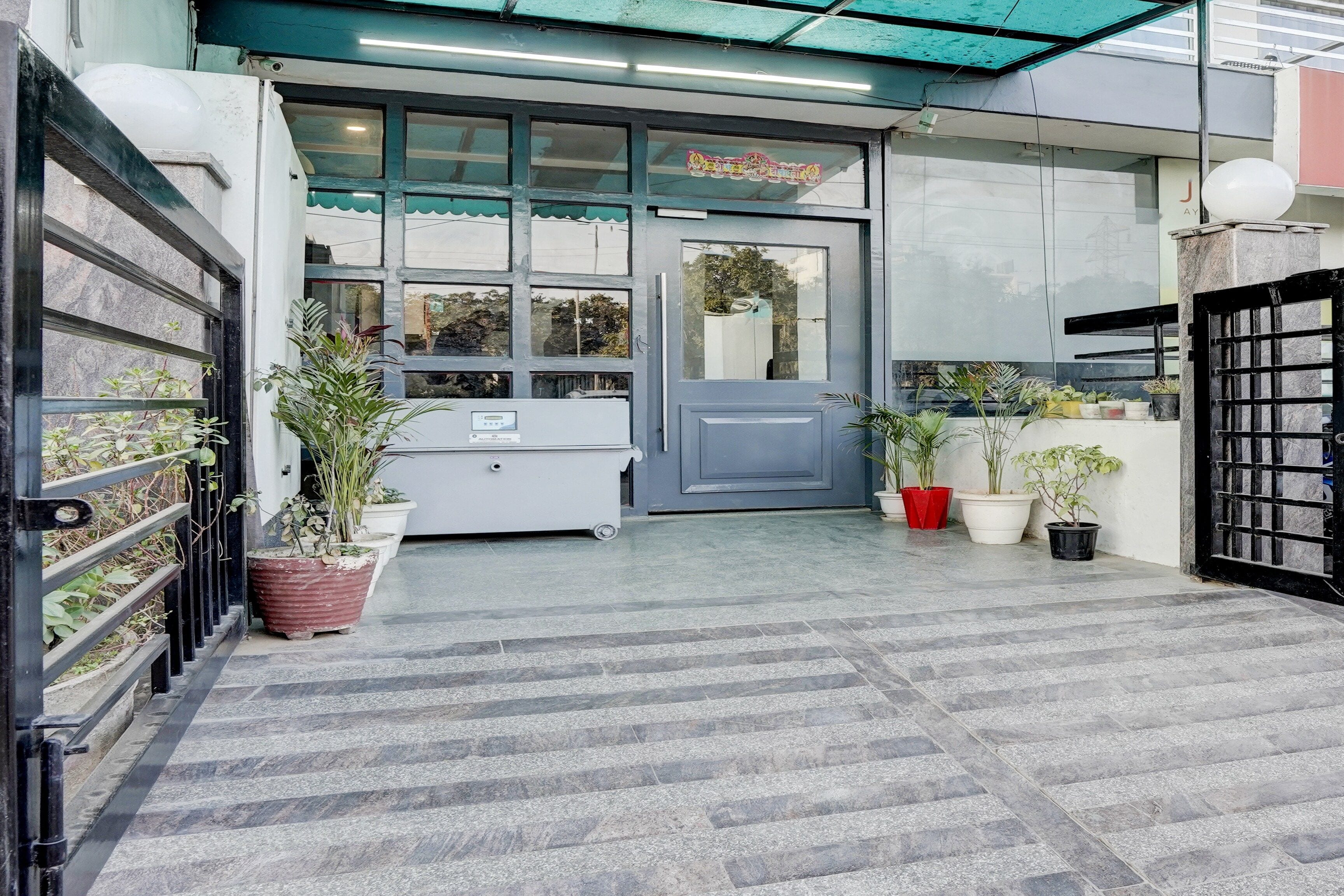Entrada de edificio con puerta gris, plantas en macetas y cámara de seguridad.