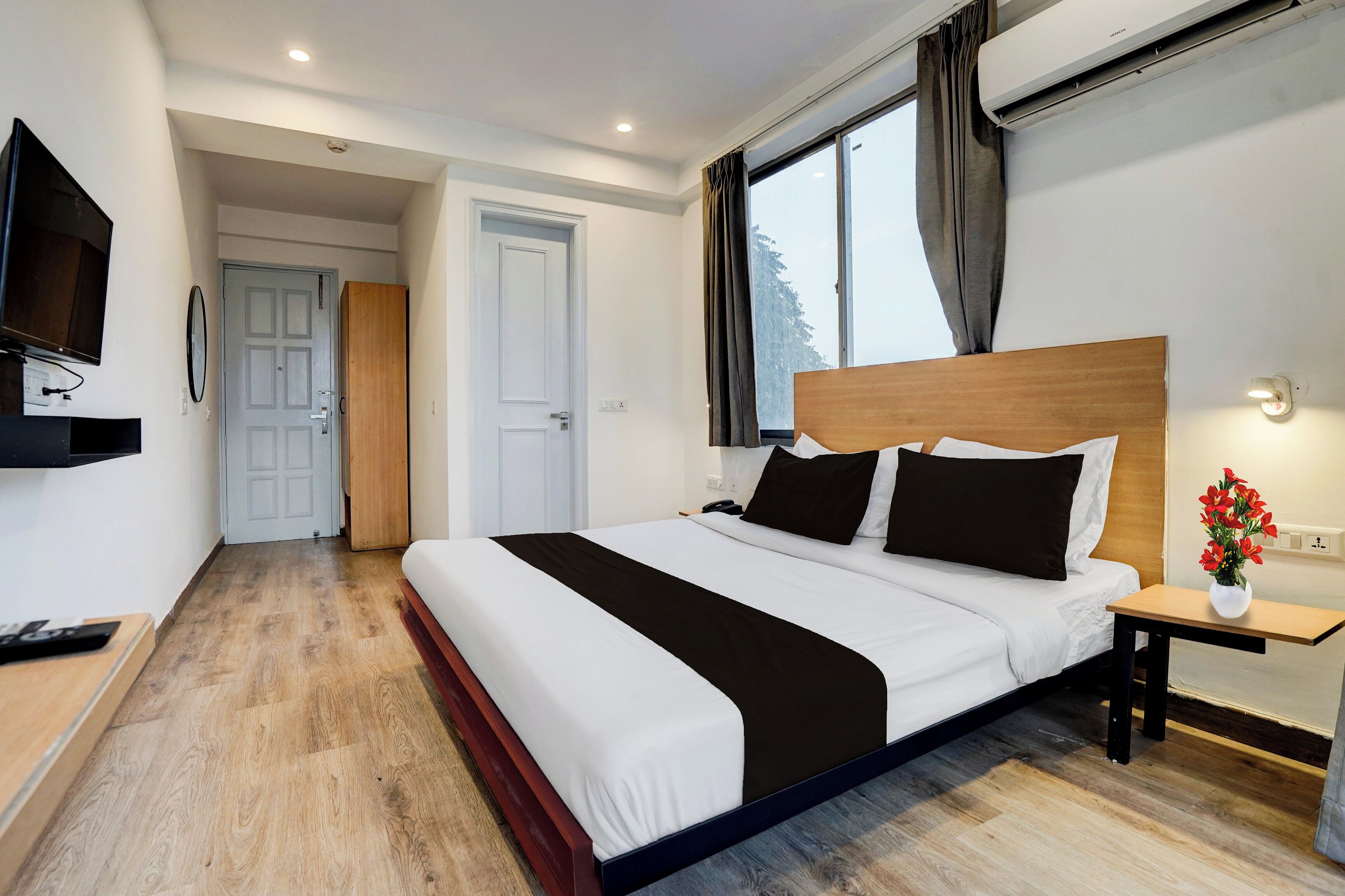 Una habitación de hotel con una cama grande, un televisor, una mesita de noche con un jarrón de flores y una ventana con cortinas.