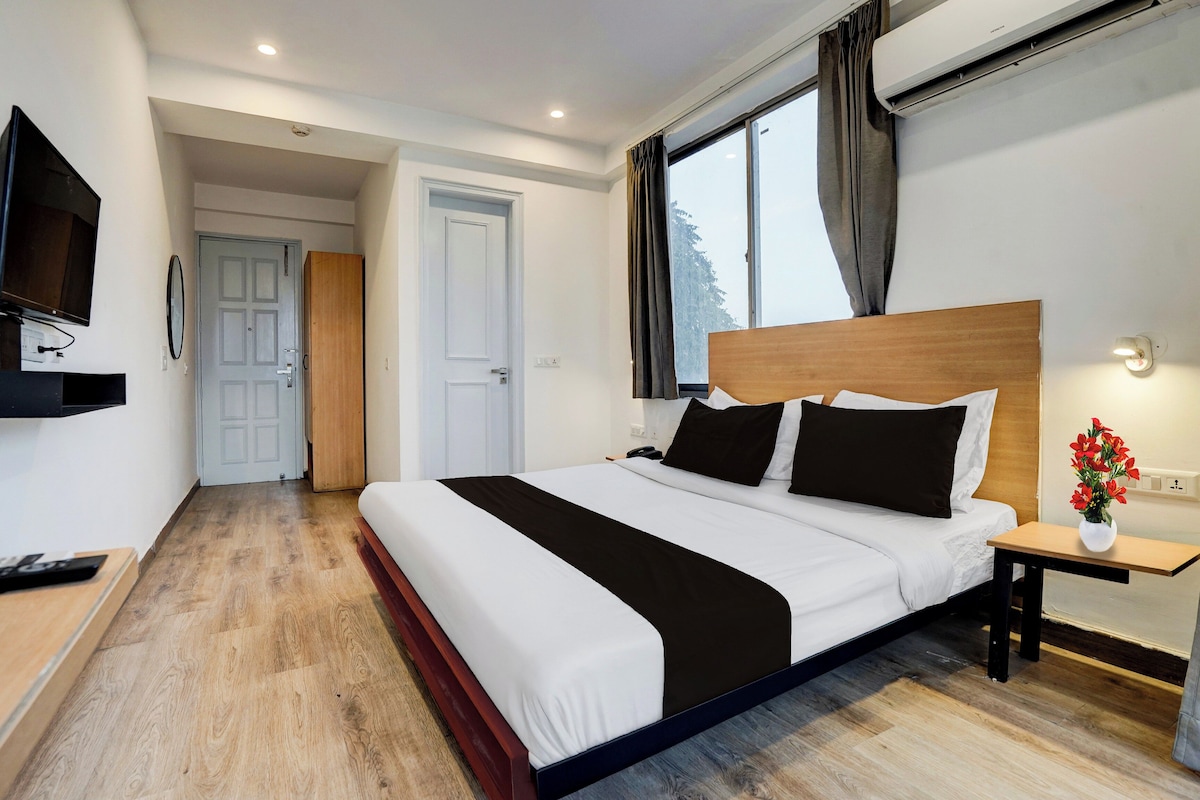 A hotel room with a large bed, a TV, a bedside table with a vase of flowers, and a window with curtains.