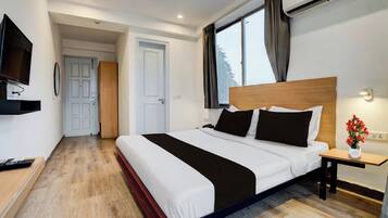 Una habitación de hotel con una cama grande, un televisor, una mesita de noche con un jarrón de flores y una ventana con cortinas.