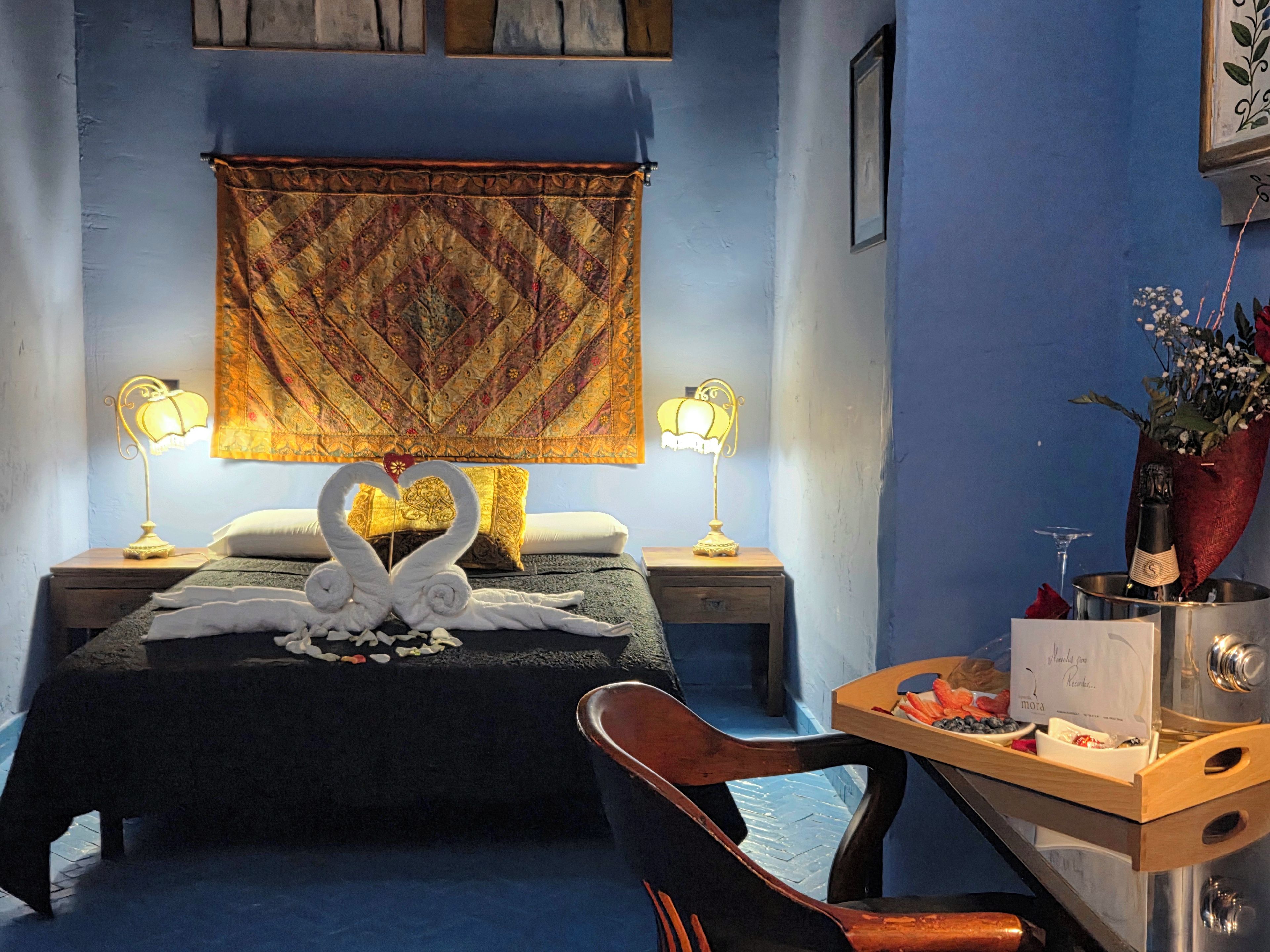 Habitación de hotel con cama, dos lámparas de noche, cabecera decorativa con forma de cisne, una mesita con una botella y flores, y una silla.