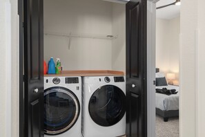 Ein Waschraum mit zwei Waschmaschinen und ein Regal für Reinigungsmittel, daneben ein Bett im angrenzenden Zimmer.