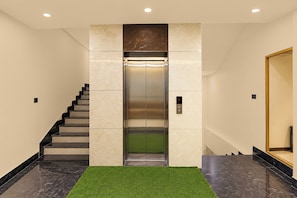 A modern elevator with a digital display, a staircase with black steps, and a green artificial grass area at the base.