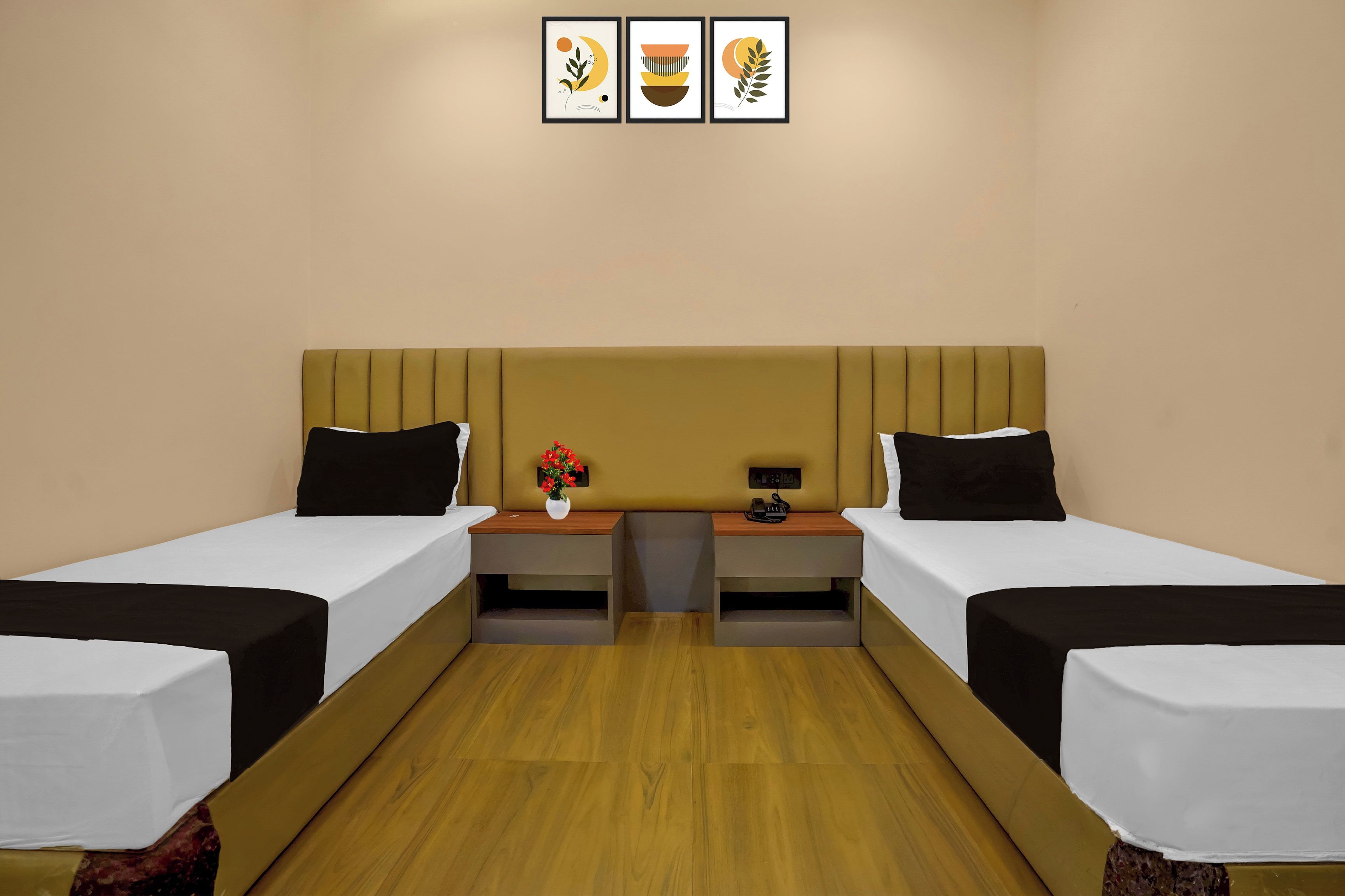 A hotel room with two beds, a wooden floor, a nightstand with a vase of flowers, and a wall-mounted power outlet.
