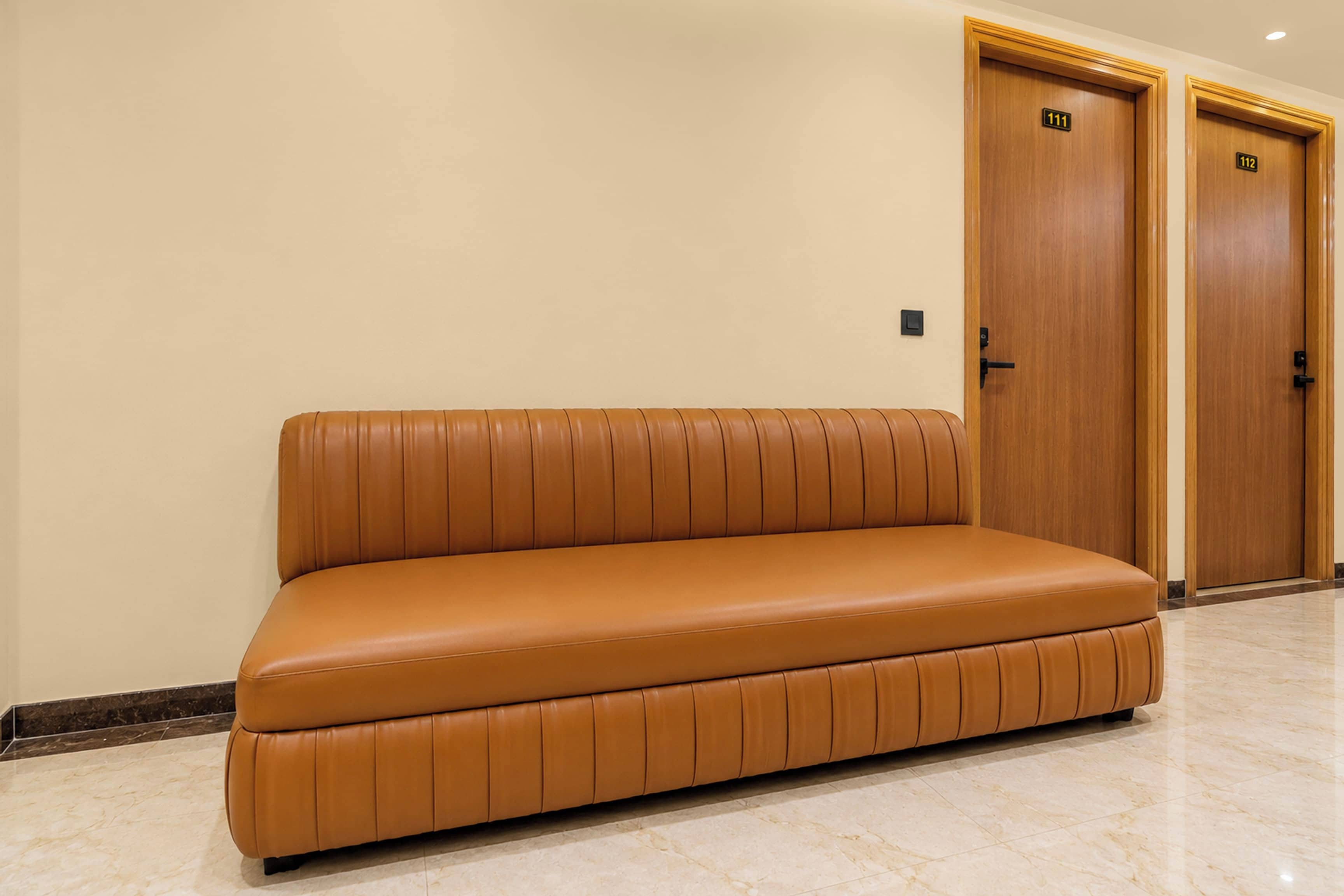 Un sofá de cuero color bronce en un pasillo con suelo de mármol y puertas de madera con las etiquetas 111 y 112.