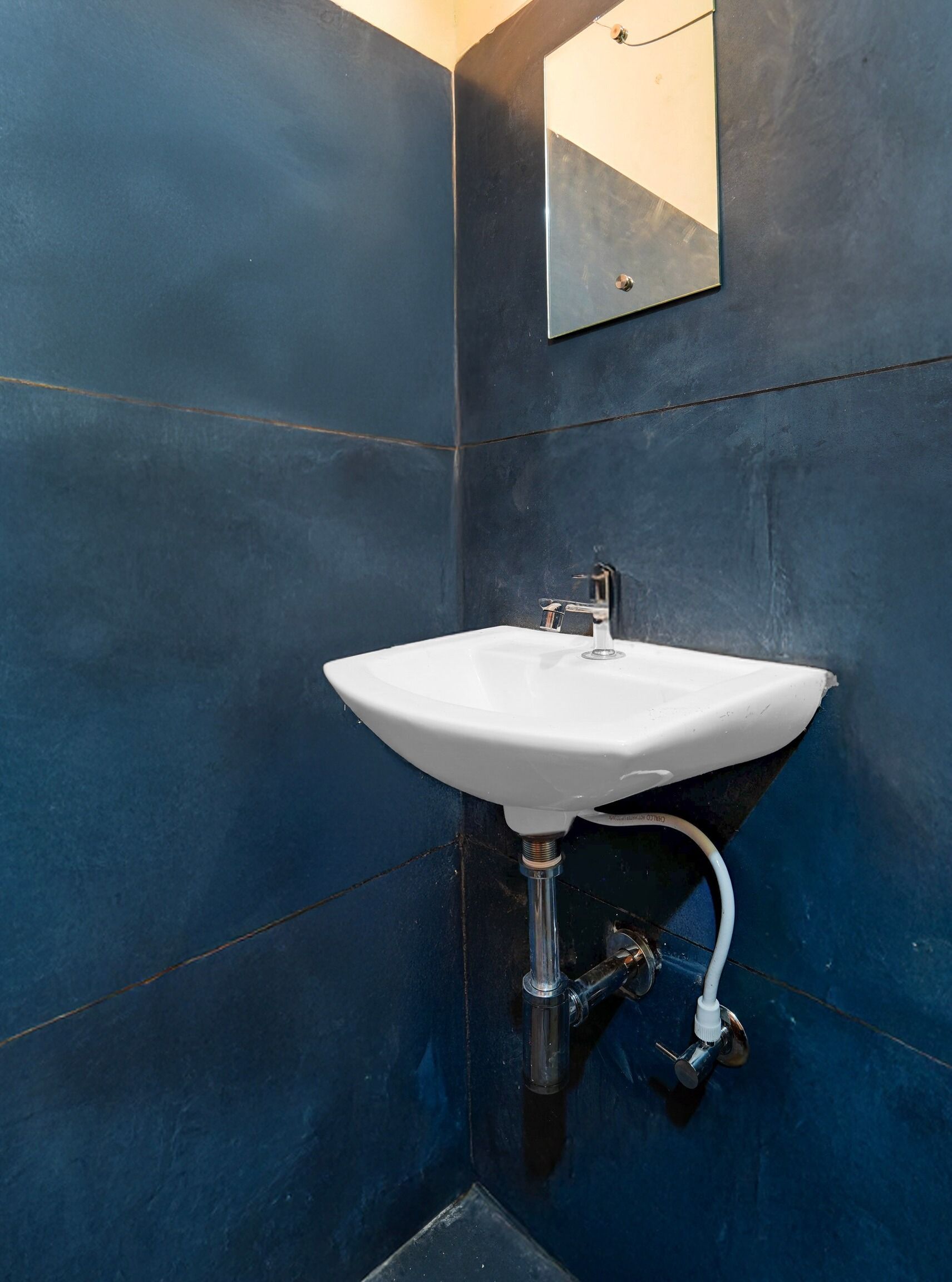 Un baño con un lavamanos blanco montado en una pared azul oscuro, un espejo sobre el lavamanos y una instalación sanitaria visible.