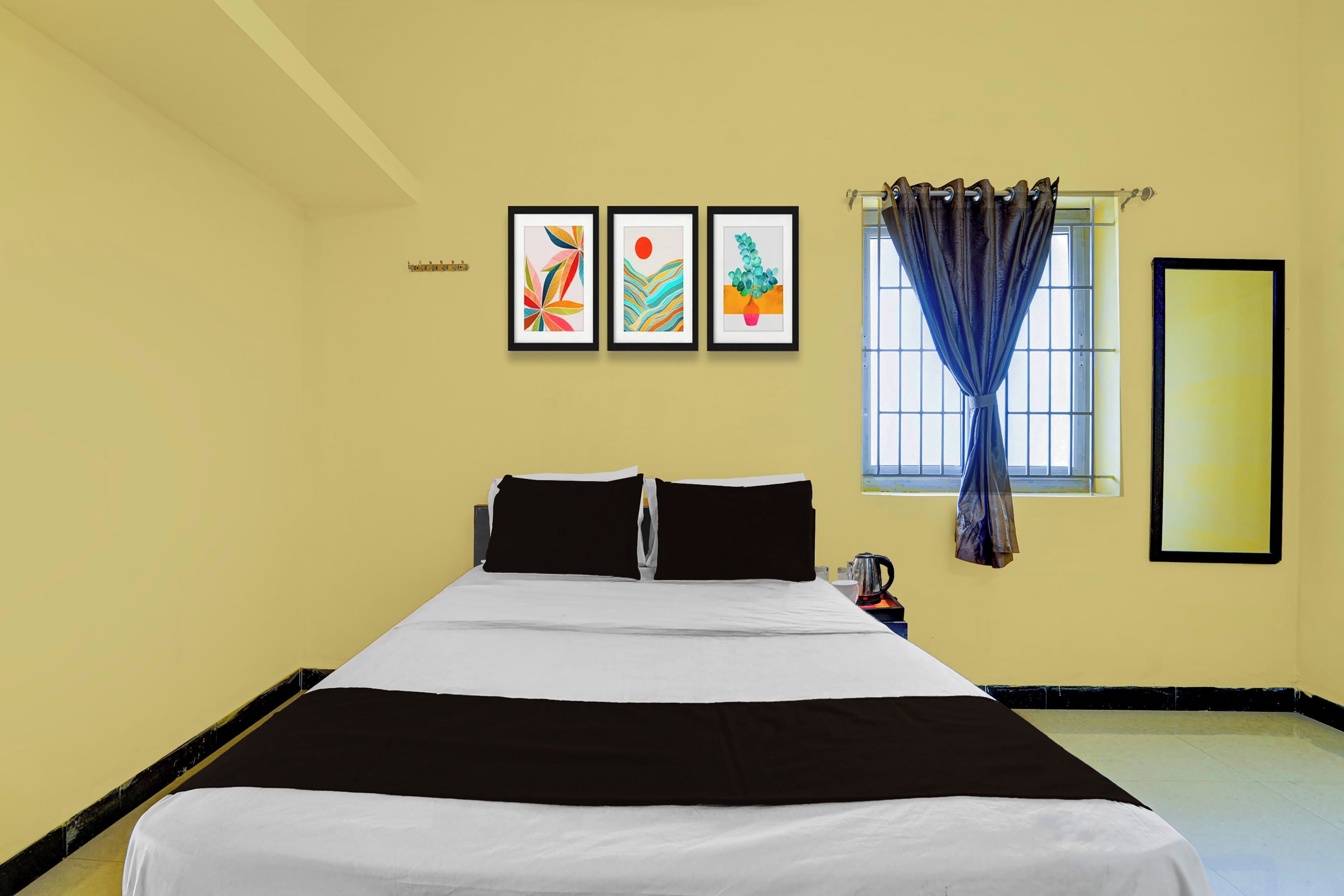 A hotel room with a bed, a window with curtains, and three framed artworks on the wall.