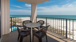 A balcony with a round table and four chairs, overlooking a beach with a pool and a clear sky. - Phoenix East 808 1 Bedroom Condo (Orange Beach)