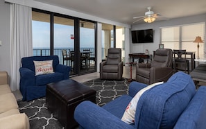 A modern living room with a blue sofa, two brown leather chairs, a dark ottoman, a flat-screen TV, and a balcony with a view of the ocean. - Phoenix Ii 2081 3 Bedroom Condo (Orange Beach)