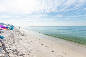 Ein Sandstrand mit Fußspuren, die zum Wasser führen, eine Reihe bunter Sonnenschirme und ein klarer Himmel.