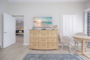 A room with a wooden cabinet, a painting of a beach scene, and a dining area with a table and chairs. - Phoenix Ii 2153 3 Bedroom Condo (Orange Beach)