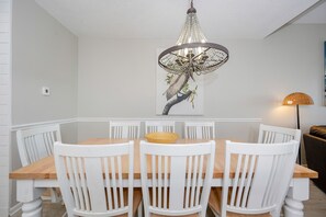A dining area with a wooden table and white chairs, a chandelier, and a wall-mounted painting. - Phoenix Ii 2153 3 Bedroom Condo (Orange Beach)
