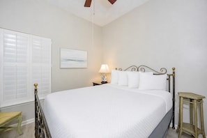 Quarto com cama, mesa de cabeceira, luminária e um banquinho pequeno.