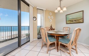 A dining area with wicker chairs and a table, a painting on the wall, and a view of the beach through a glass door. - Phoenix East 505 1 Bedroom Condo (Orange Beach)