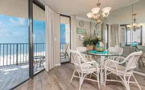 A room with a glass table surrounded by wicker chairs, a view of the beach, and a chandelier. - Phoenix East 705 1 Bedroom Condo (Orange Beach)