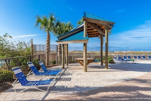 A beachside lounge area with blue lounge chairs, a wooden pergola, and palm trees. - Phoenix East 408 1 Bedroom Condo (Orange Beach)