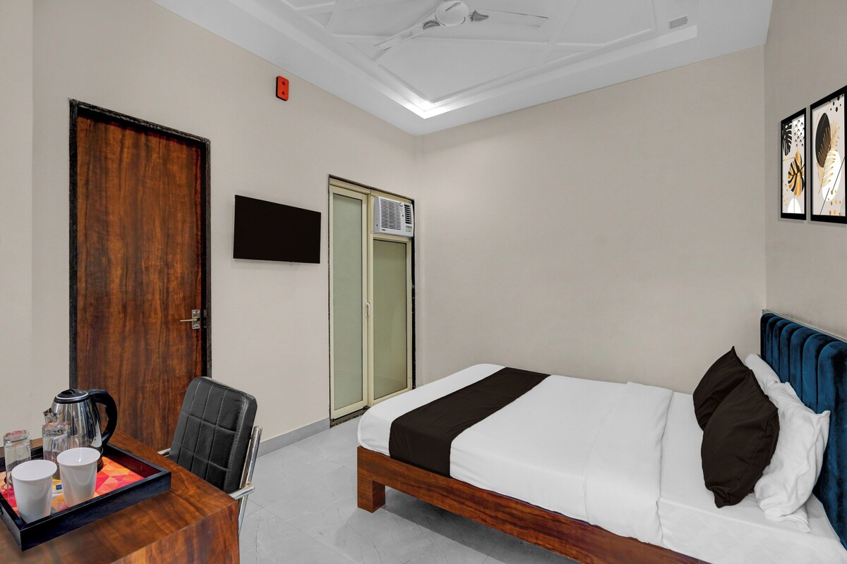 A hotel room with a bed, a desk with a kettle, cups, and a chair. There is a door, a window, and a ceiling fan.