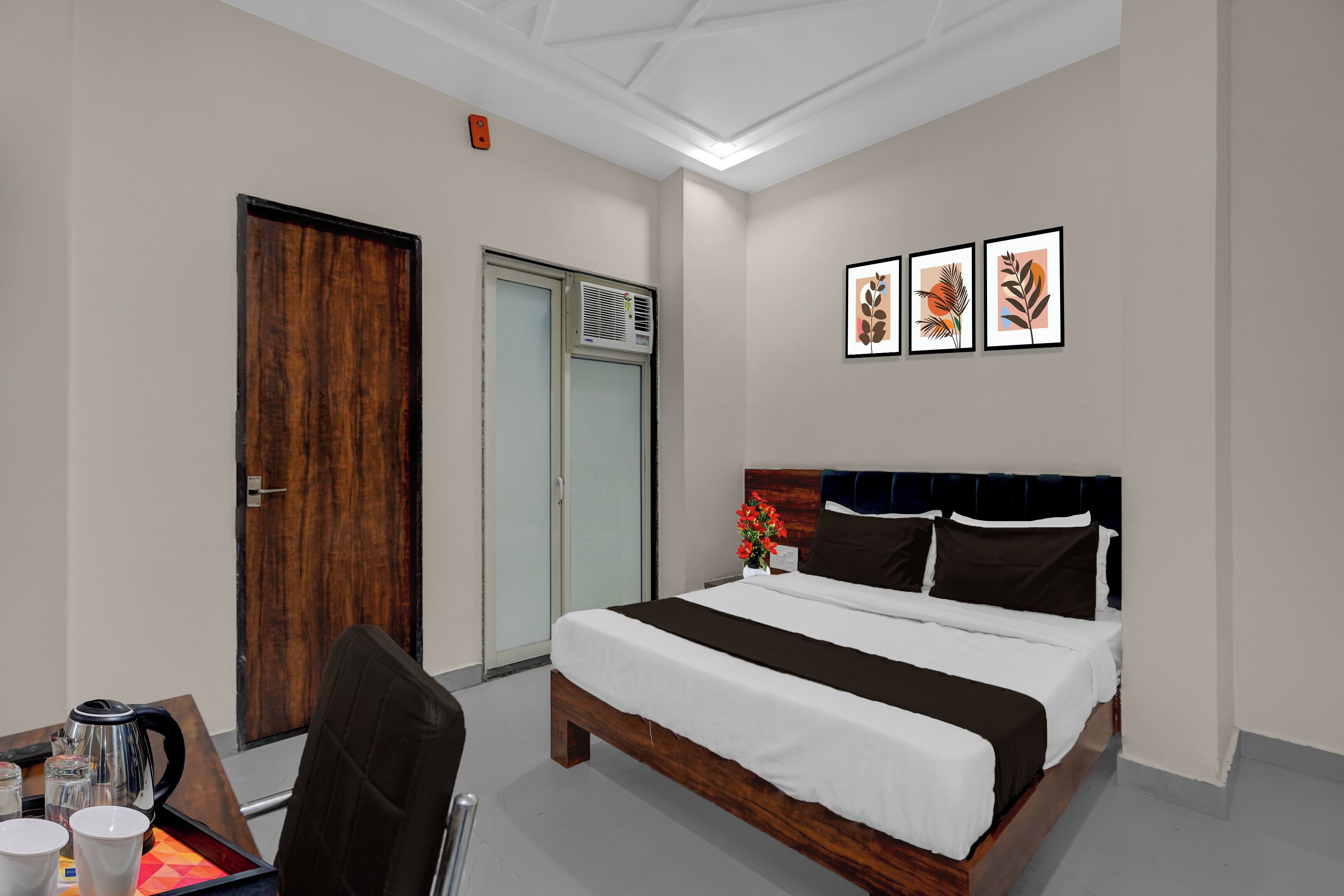 Habitación de hotel con cama, escritorio, silla, cafetera y un cuadro de una planta en la pared.