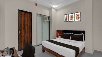 Habitación de hotel con cama, escritorio, silla, cafetera y un cuadro de una planta en la pared.