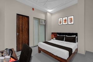 A hotel room with a bed, a desk, a chair, a coffee maker, and a picture of a plant on the wall.