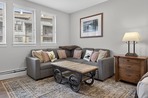 A living room with a gray sectional sofa, a wooden coffee table with wheels, a patterned rug, a framed picture on the wall, and a lamp on a wooden side table. - Premium Unit 4571 - Studio - Founders Pointe Condo (Winter Park)