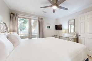Quarto de hotel com cama, ventoinha no teto, televisão, mesa de cabeceira, espelho e janela com cortinas.