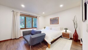 A bedroom with a bed, a chair, a nightstand, a lamp, a vase with branches, and a painting on the wall. - Meadow Ridge #8 3 Bedroom Condo (Mammoth Lakes)