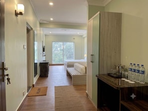 A hotel room with a bed, a bedside table, a nightstand, a wardrobe, a desk with water bottles, and a view of the outdoors.