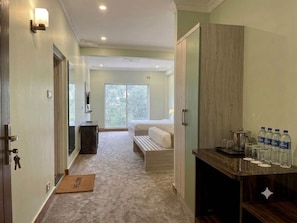 A hotel room with a bed, a bedside table with water bottles, a desk with a computer, and a view of greenery outside.
