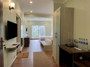 A hotel room with a bed, a TV, a desk with a computer, and a window with a view of trees.
