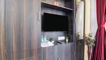 Un mueble de madera oscura con un televisor de pantalla plana, un estante pequeño con botellas de agua, vasos y un control remoto, y una planta en maceta.