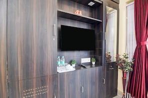 A dark wood entertainment unit with a flat-screen TV, a small shelf with water bottles, glasses, and a remote control, and a potted plant. - Super Collection O Jyotirindra Nandi (Kolkata)