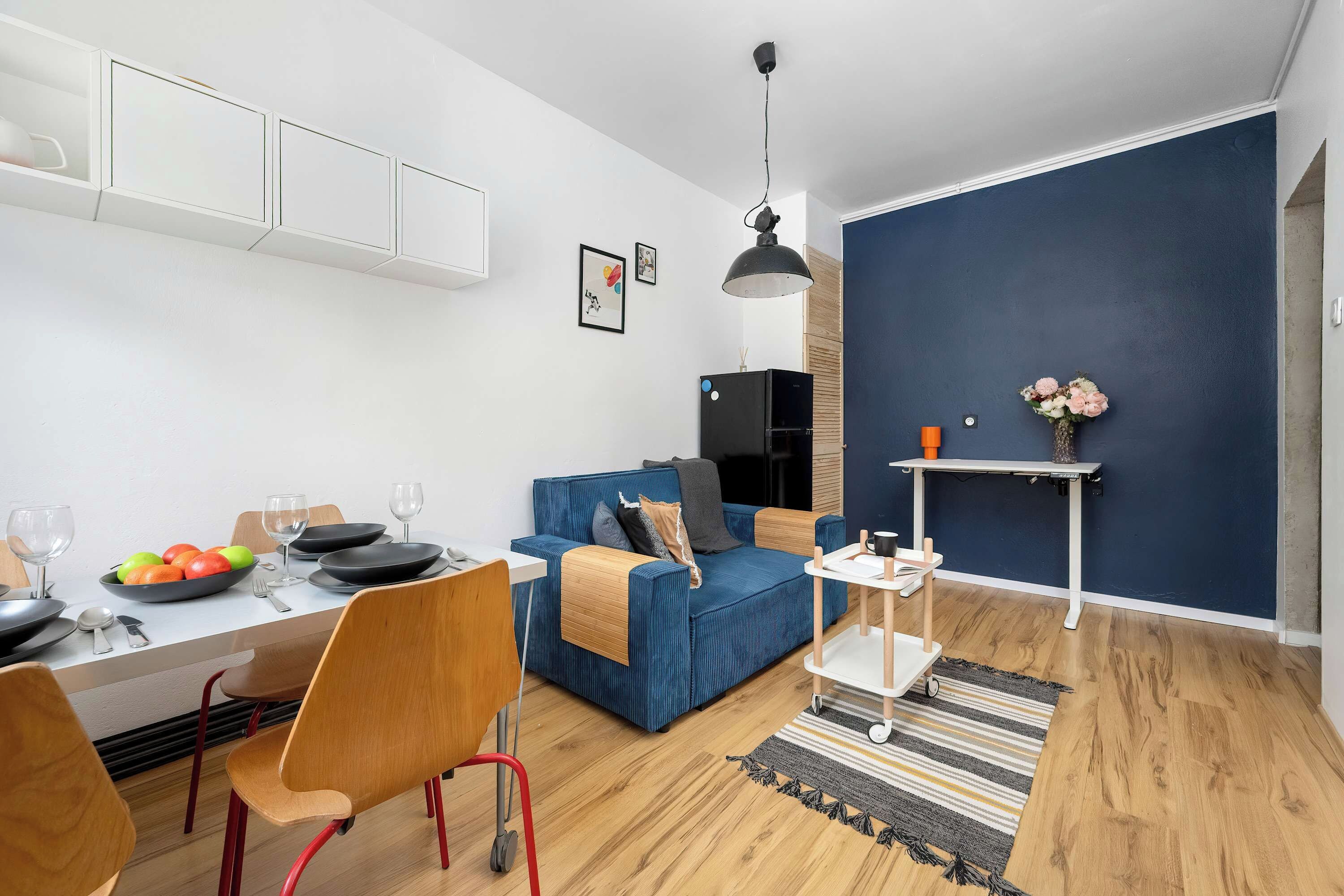 Ein modernes Wohnzimmer mit einem blauen Sofa, einem Esstisch mit Stühlen, einem kleinen weißen Tisch und einer blauen Akzentwand.